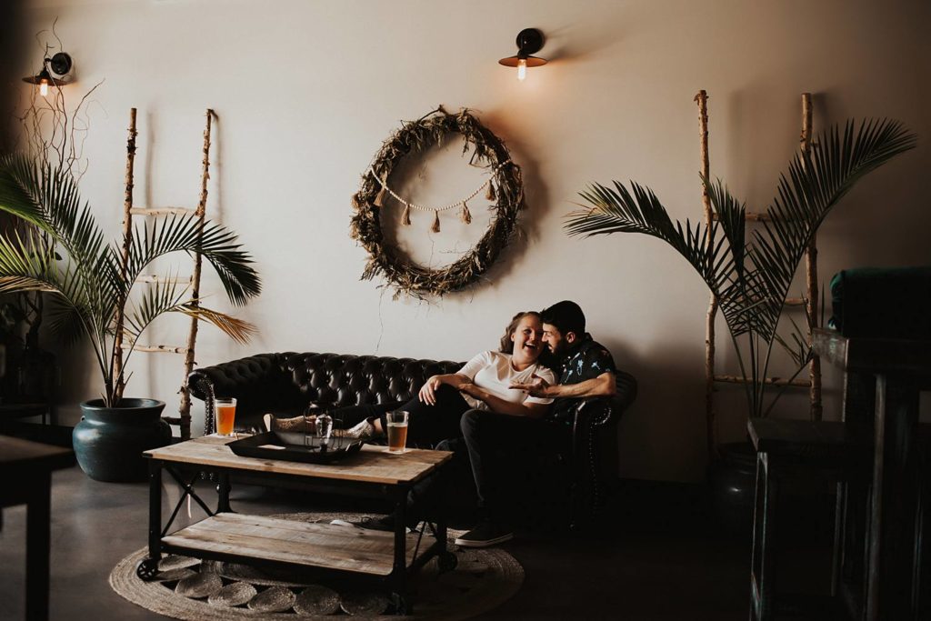 Couple sitting on couch laughing during engagement session at brewery
