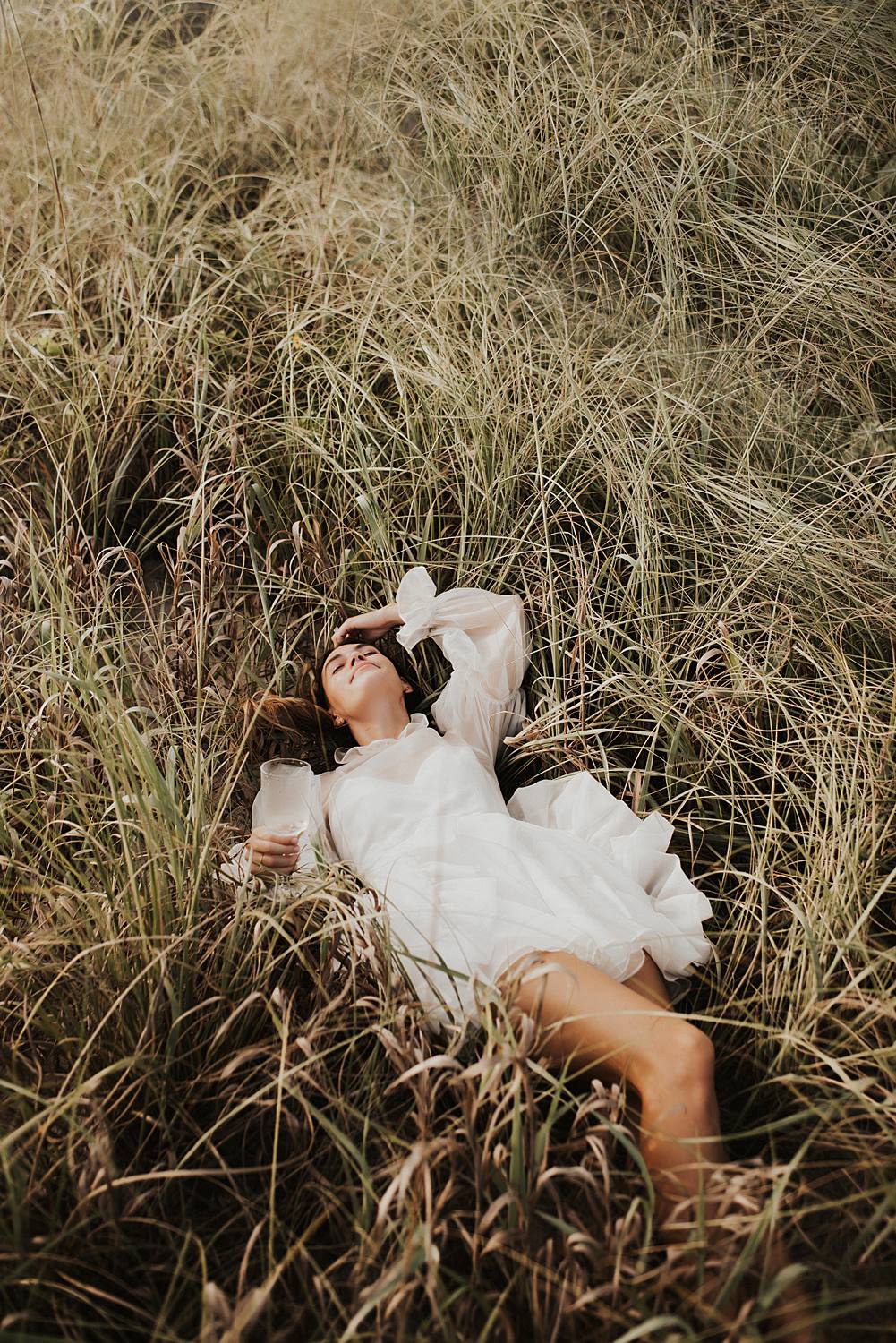 Bride laying in tall beach grass with glass of champagne