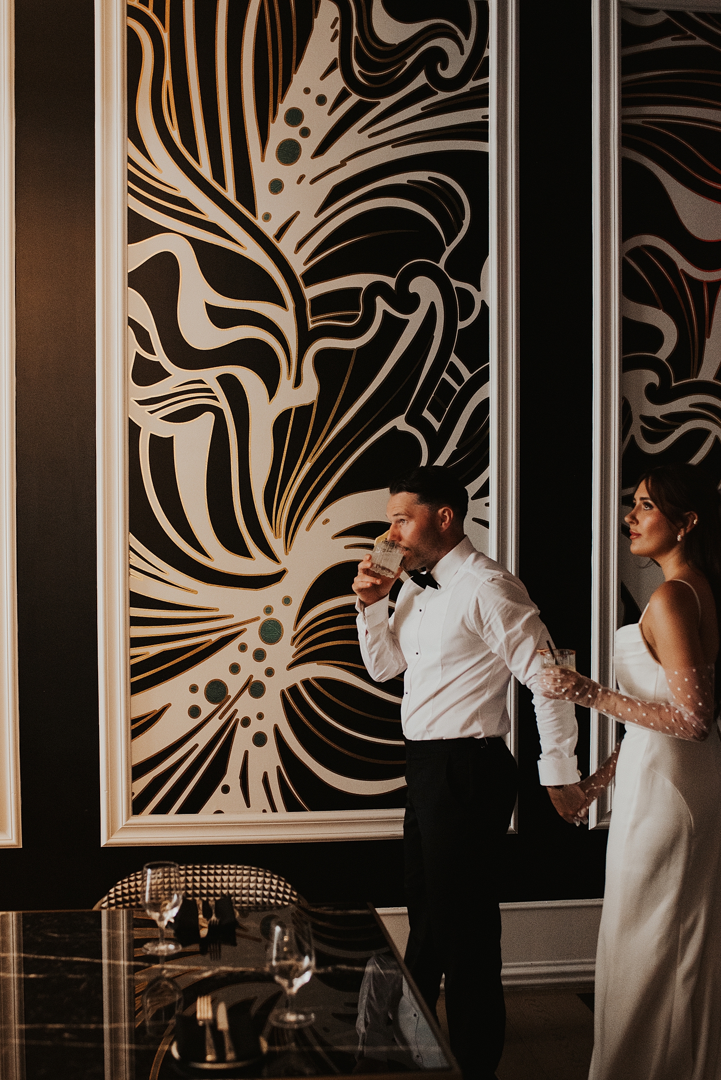 Groom leads bride through a room with large paintings on the wall at the Grand Bohemian as he sips a cocktail after their Orlando elopement