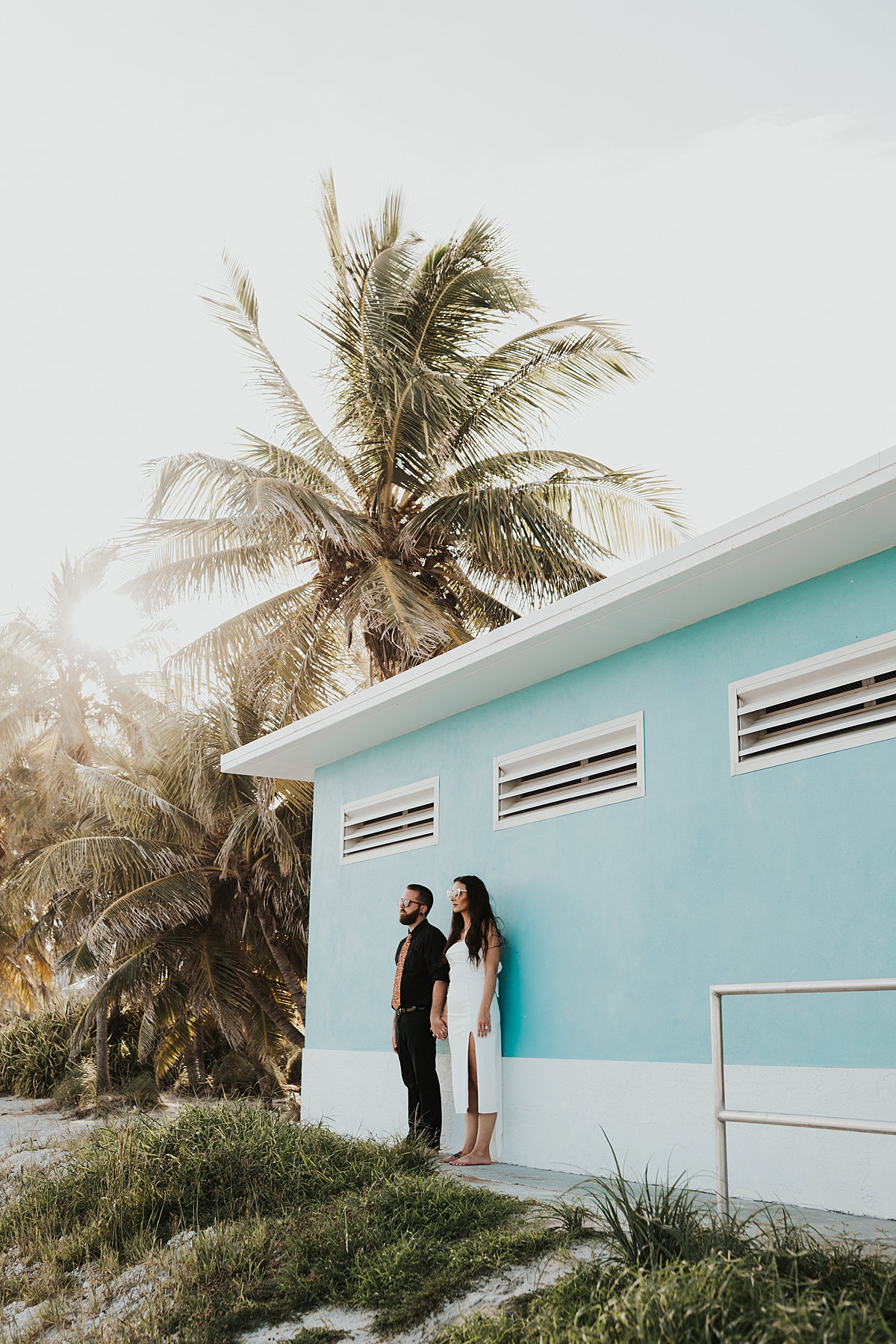 Bride in heart shaped glasses with mid length wedding dress and groom in sunglasses standing up against a bright blue wall with palm trees in the back after their Key West elopement on Smathers Beach