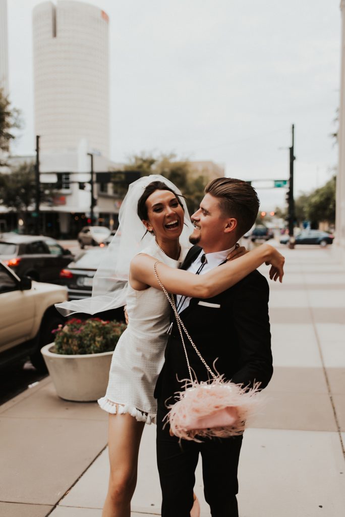 Bride in short white dress and veil hugging groom from behind on downtown tampa street