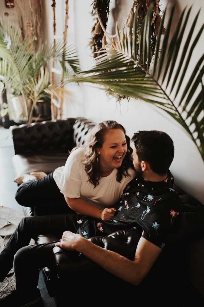 Couple sitting on couch laughing during engagement session at brewery