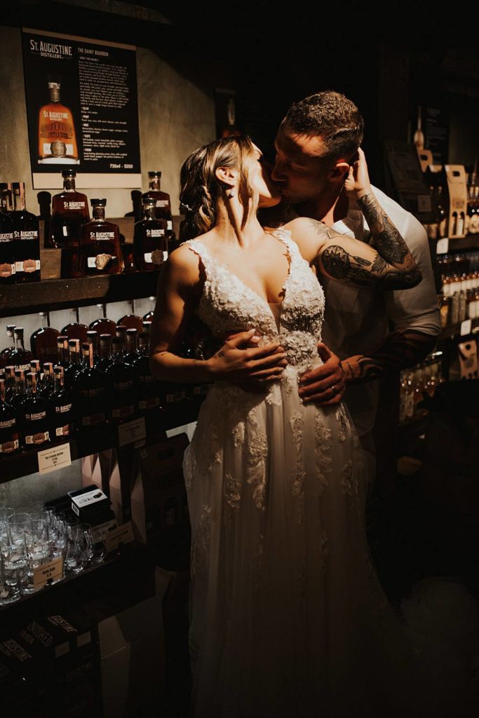 Bride and groom kissing in front of whiskey bottles at Saint Augustine Distillery
