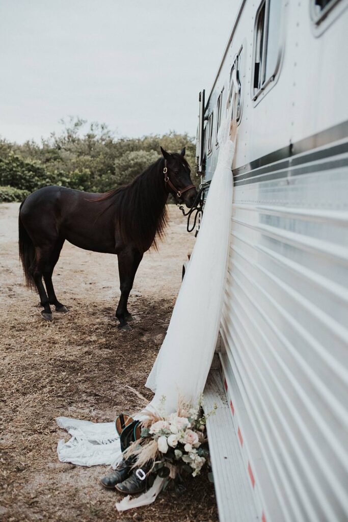Brides wedding gown hung up with boots and flowers on the ground with her horse in the background