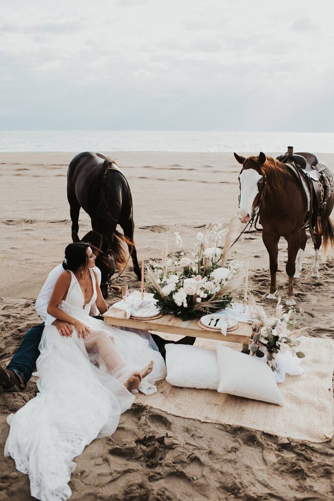 Luxury picnic on Florida beach with pillow seats and floral cake meadow with bride and groom sitting on the ground with their horses