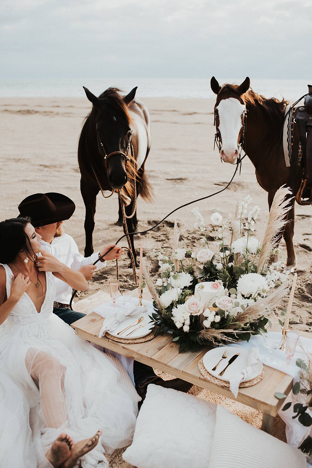 Luxury picnic on Florida beach with pillow seats and floral cake meadow with bride and groom sitting on the ground with their horses