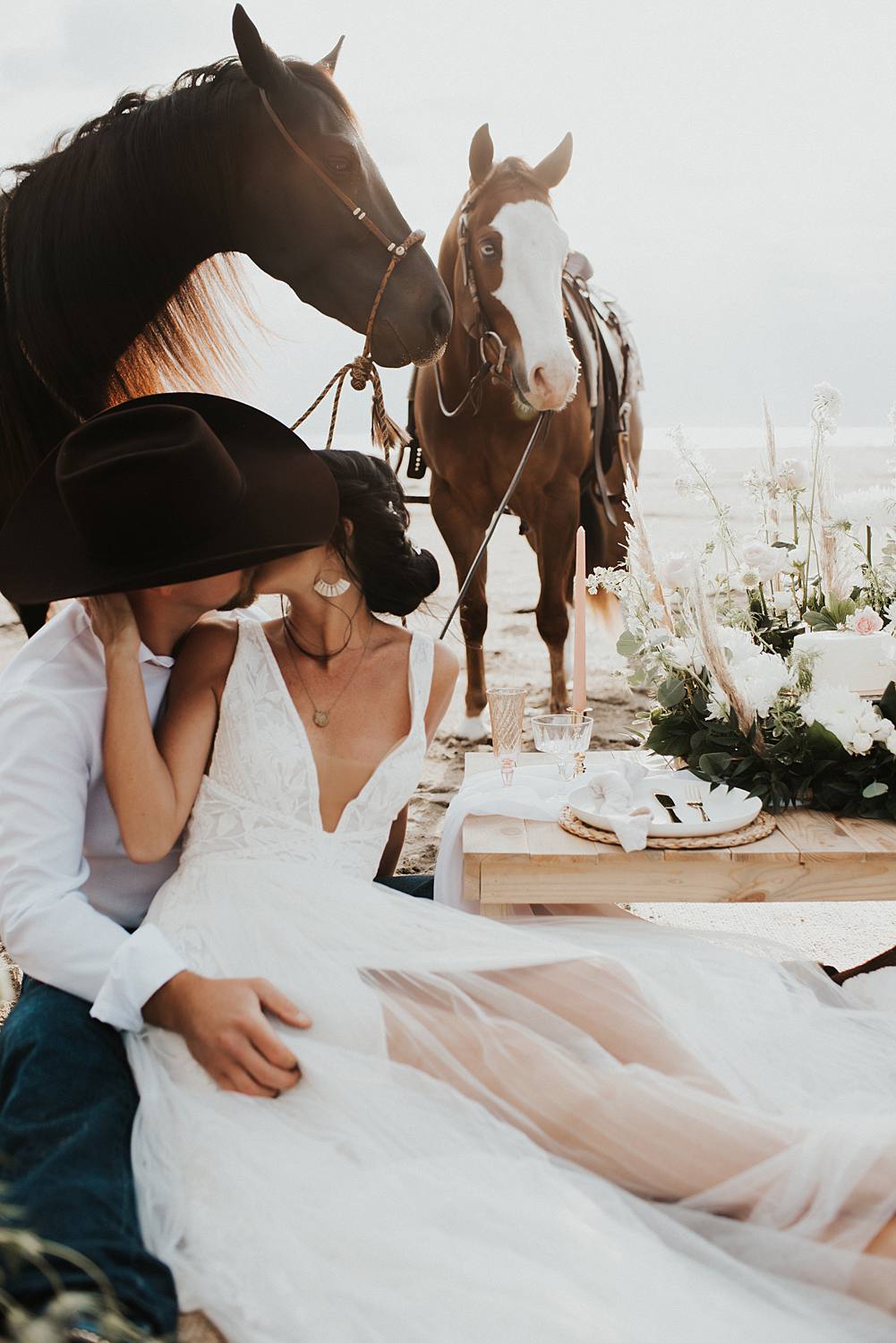 Luxury picnic on Florida beach with pillow seats and floral cake meadow with bride and groom sitting on the ground with their horses