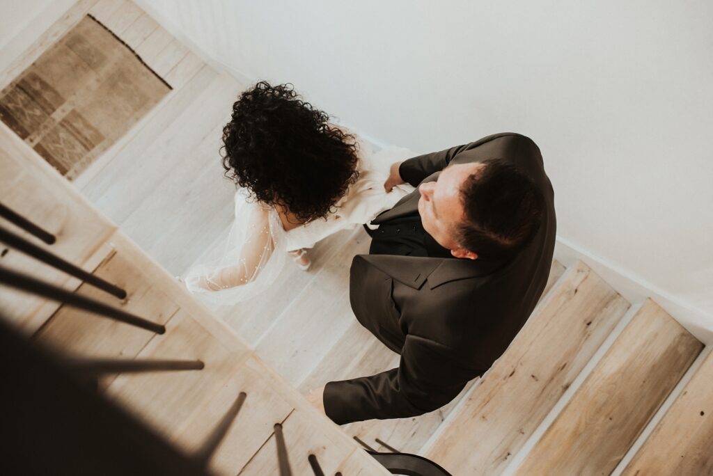 Groom holding bride's dress train as they walk down the stairs