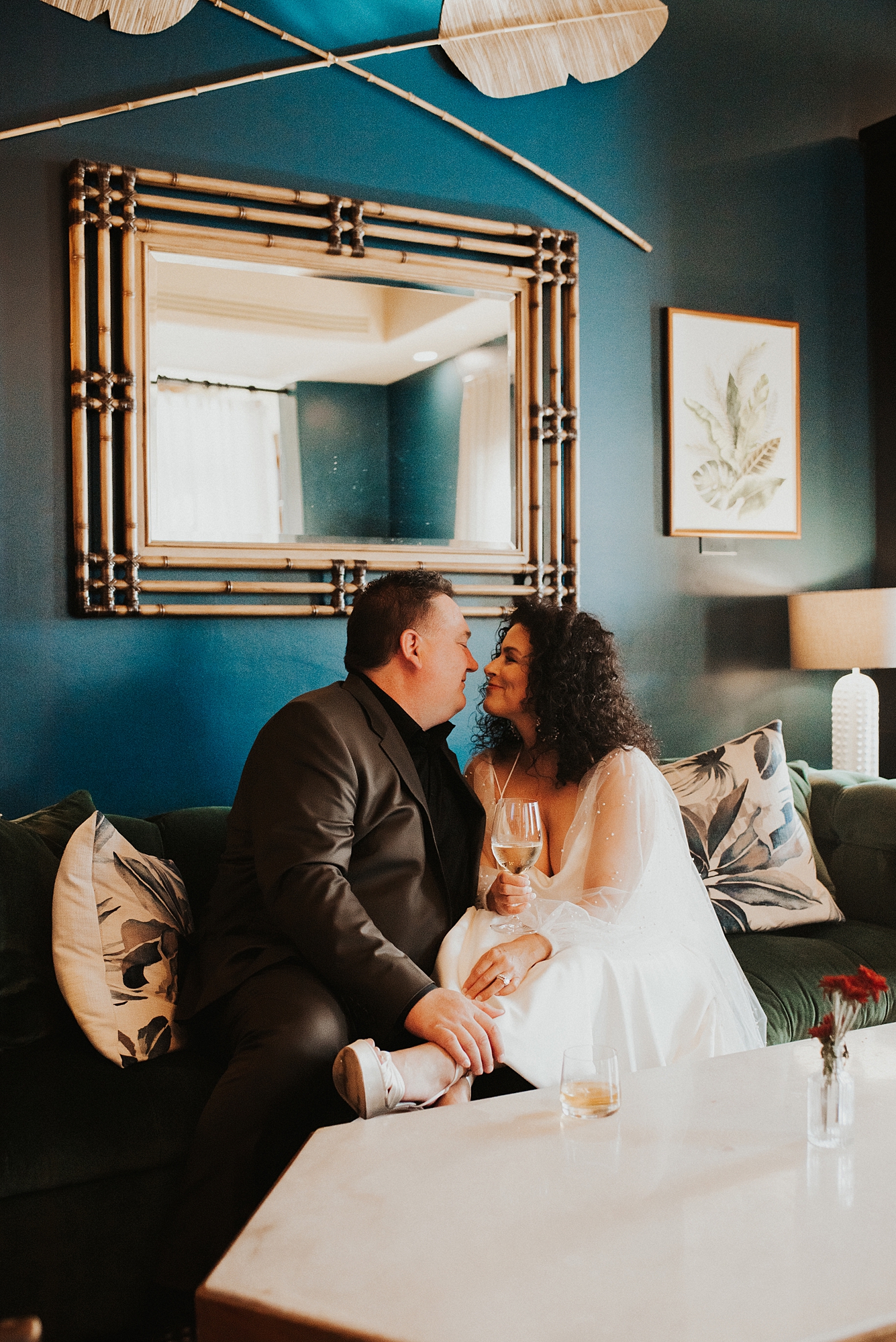 Bride and groom sitting on a couch in front of a blue wall sharing a cocktail